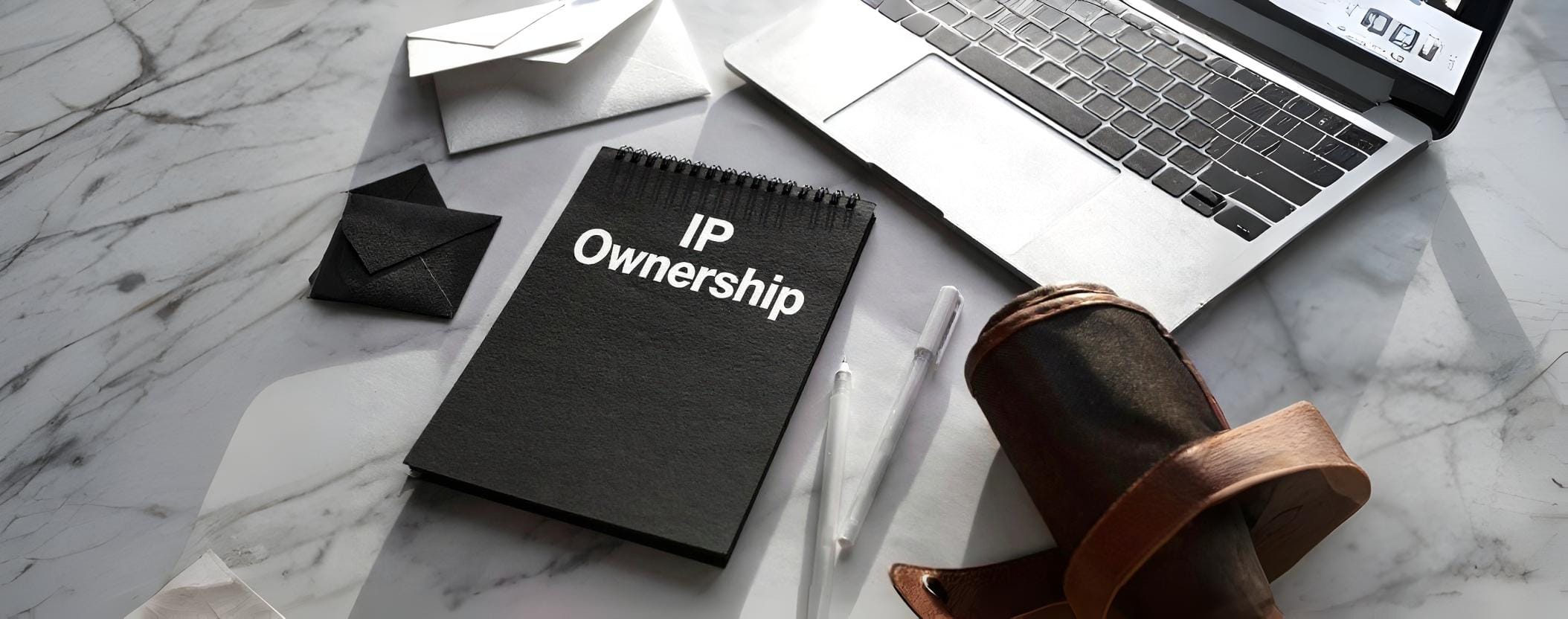 Notebook with the words ‘IP Ownership’ on a marble desk beside a laptop and documents, symbolizing legal protection of intellectual property.