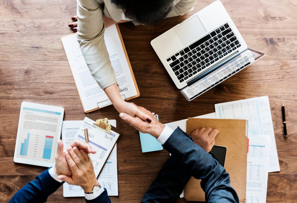 Overhead shot of two people shaking hands over a table with documents and laptop, representing a capital raise agreement or financing deal.