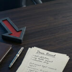 Red downward arrow on dark walnut desk beside leather portfolio and corporate documents, representing startup down round valuation drop and founder equity protection